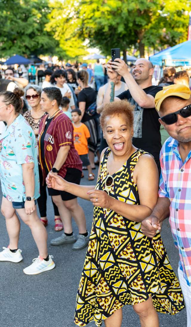 Diverse group of people dancing and smiling at a Fourth Friday event in downtown Fayetteville, NC, with vendor tents and summer sunlight in the background.