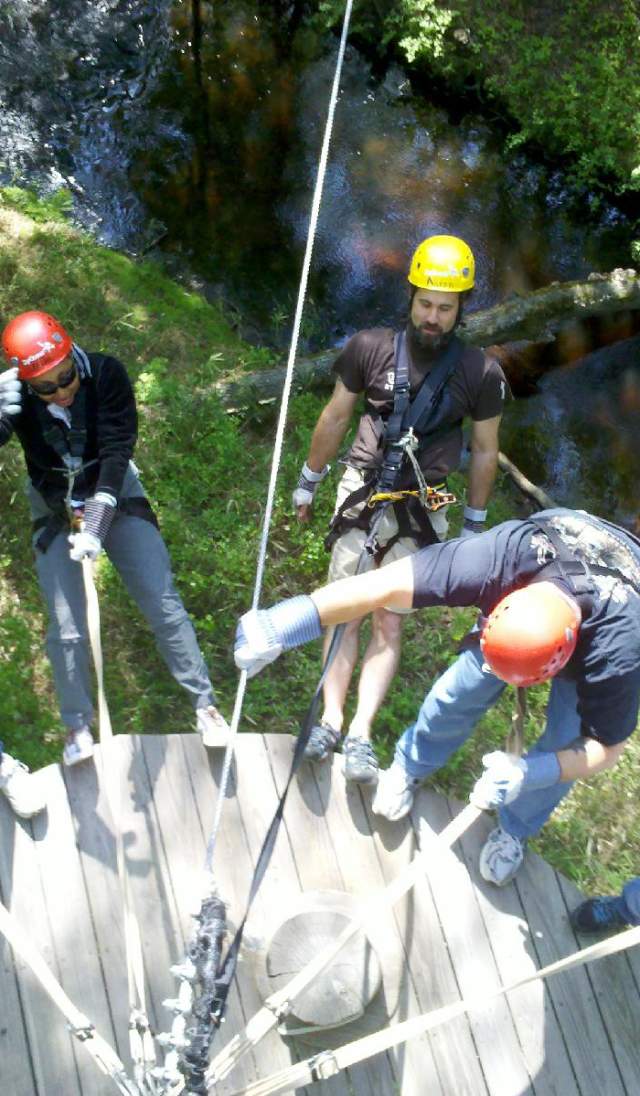 Group of people wearing helmets and safety harnesses standing on a zipline platform at ZipQuest, surrounded by trees and water below.