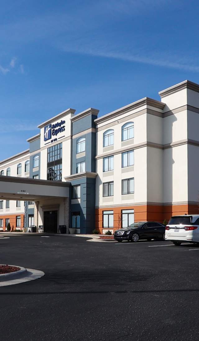 Exterior view of a multi-story hotel with a covered entrance drive, parked cars in the lot, landscaped grounds, and a clear blue sky.