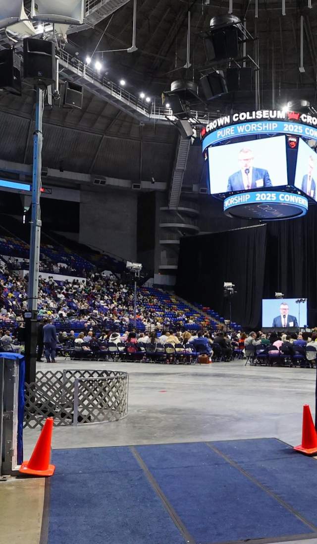 Wide view of a Jehovah’s Witness convention inside Crown Coliseum in Fayetteville, North Carolina, showing attendees seated around the arena floor with a central stage and overhead screens.