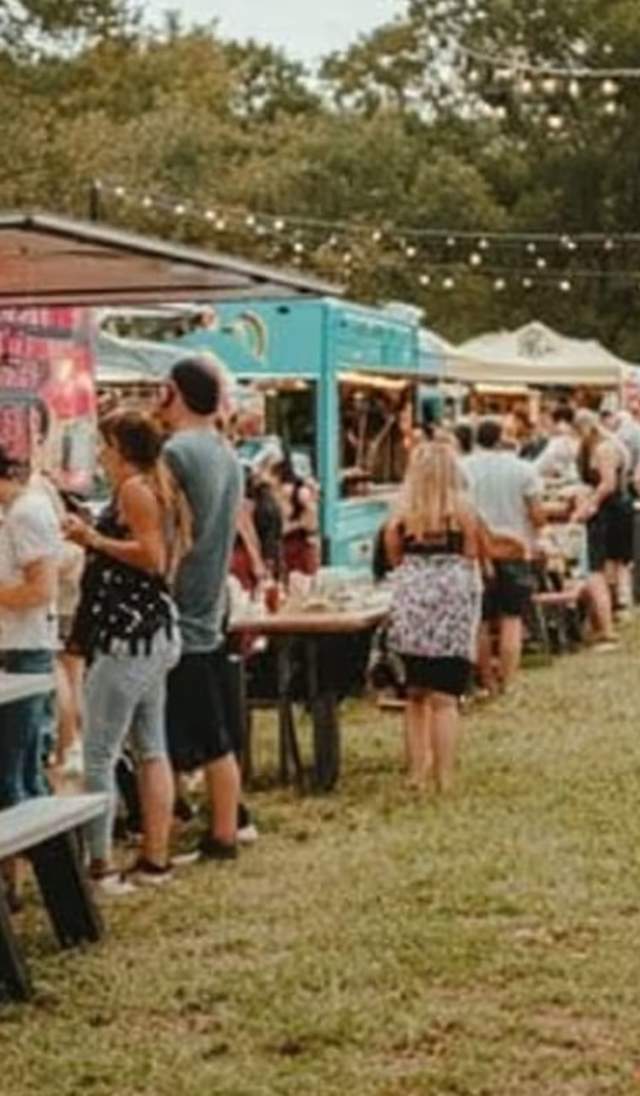 People dining and socializing at an outdoor food truck festival with picnic tables and string lights overhead