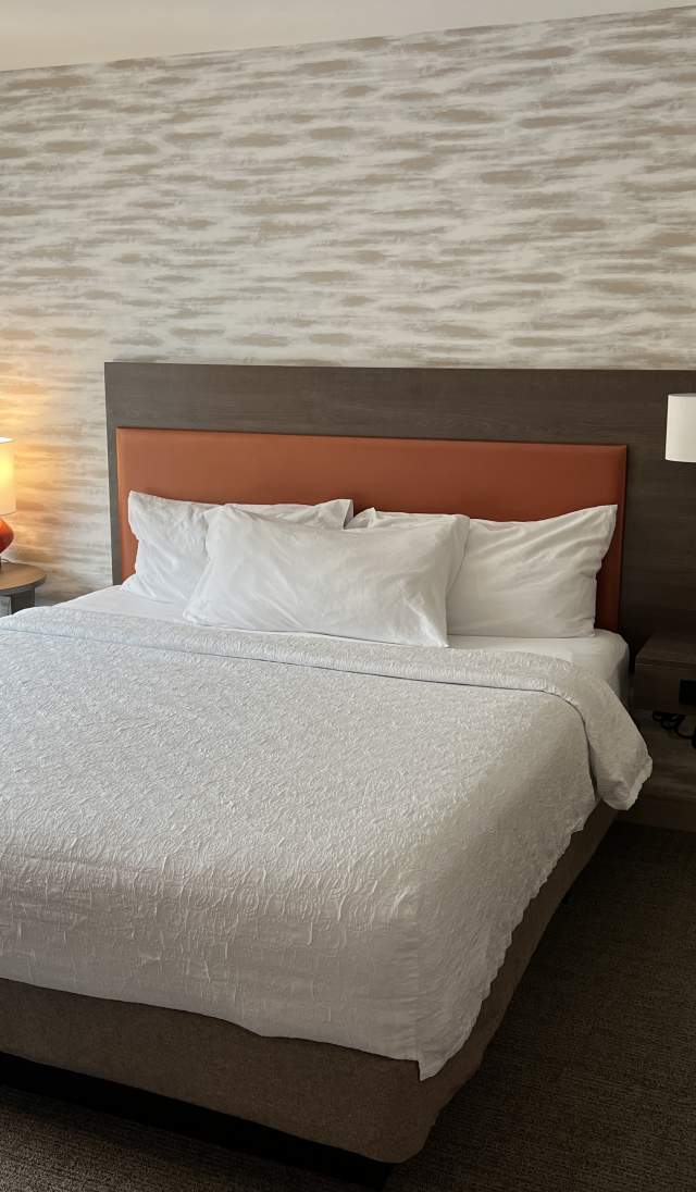 Modern hotel room with a neatly made bed, white bedding, wood headboard, bedside lamps, textured accent wall, and a window providing natural light.