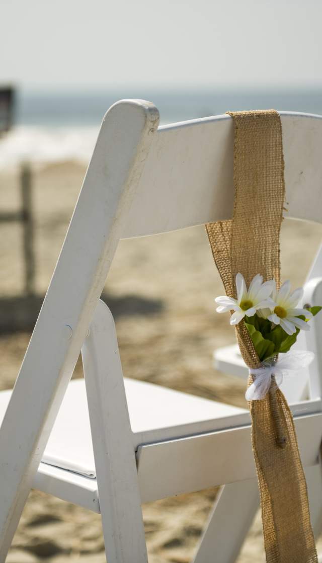 A white chair on a sandy beach is decorated with burlap and small white flowers, with a serene ocean backdrop.
