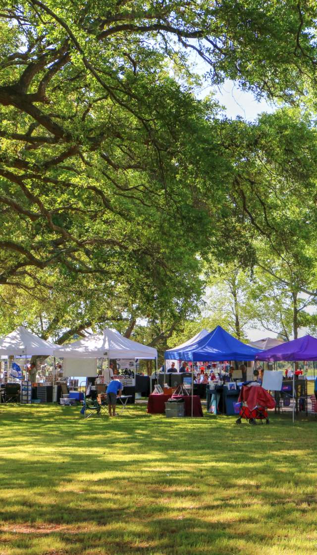 Visitors explore a vibrant outdoor market in Sunset Beach, NC, with colorful vendor tents under shady trees on a bright, sunny day.