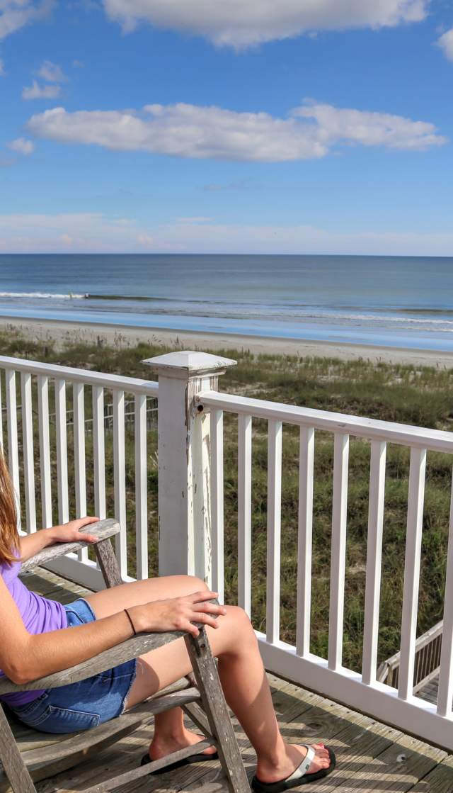Person sitting on the upper deck of an oceanfront vacation rental in the Brunswick Islands, overlooking sandy dunes and gentle ocean waves under a partly cloudy sky.