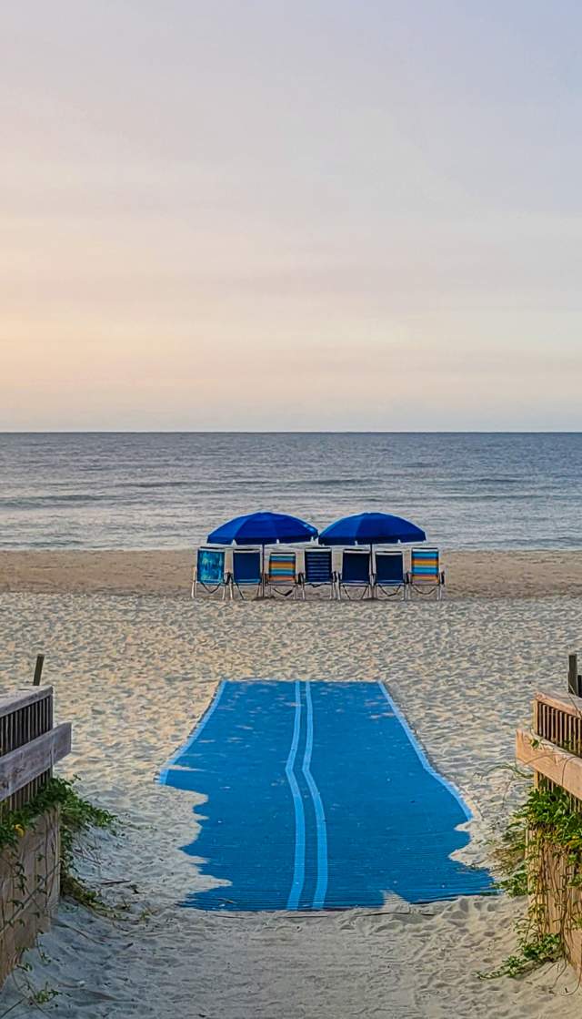 a wooden walkway and ADA beach mat leading out onto a beach with chairs and umbrellas in view