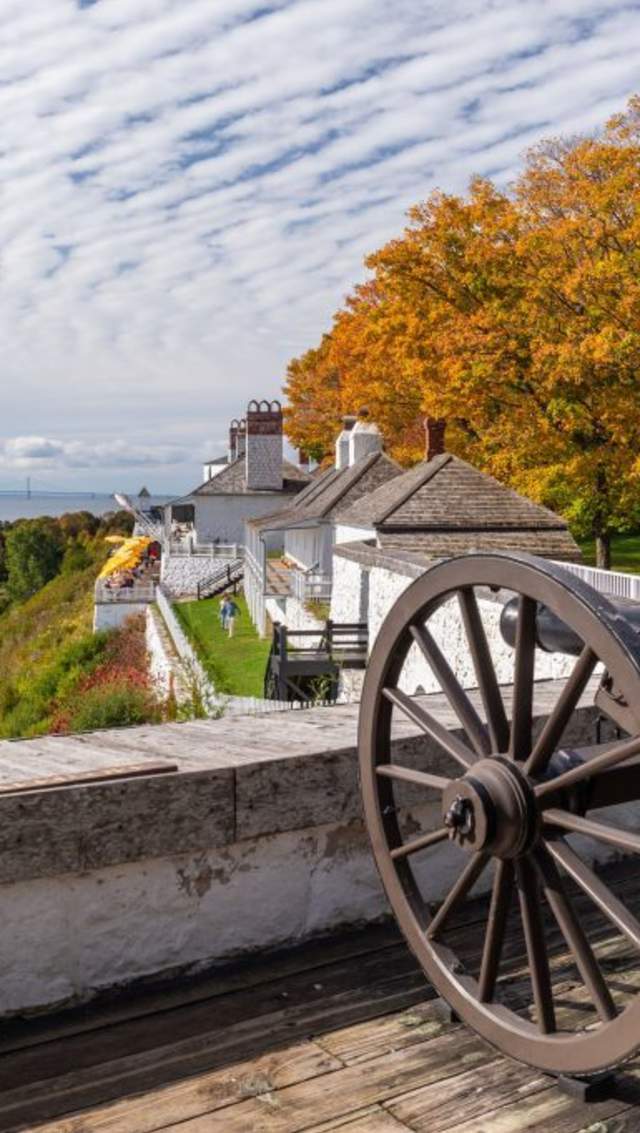 A Fort Mackinac cannon points out over town amid fall colors and blue water with the Mackinac Bridge in background