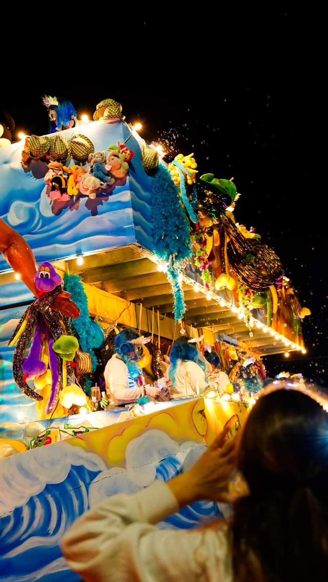 A Carnival float in the Krewe of Eve parade rolls through Mandeville, surrounded by a crowd with arms raised to catch beads.