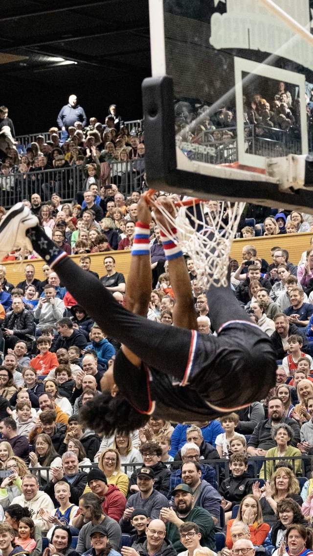 A Harlem Globetrotter player hanging off a basketball hoop with a large crowd watching at the Bournemouth International Centre in Dorset