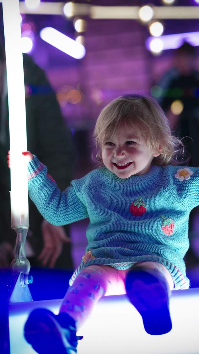 A little girl enjoys playing on an illuminated swing, with neon lights in the background