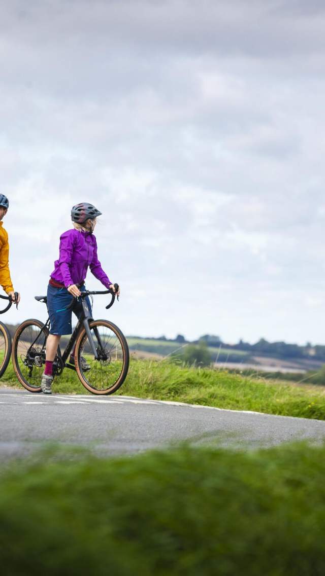 A couple cycling in the Lincolnshire Wolds on a cloudy day. They are both stopped and looking to cross a road.