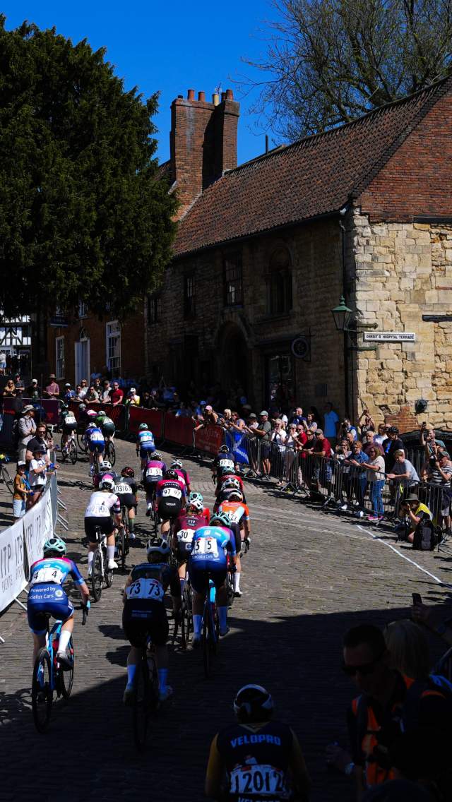 A group of cyclists compete in the Men's Tour of Britain race in Lincolnshire, with spectators watching from behind crowd barriers on either side of the route.