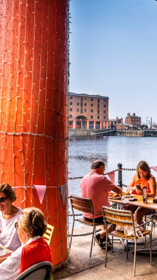 People sitting at tables alongside the Royal Albert Dock in the sunshine