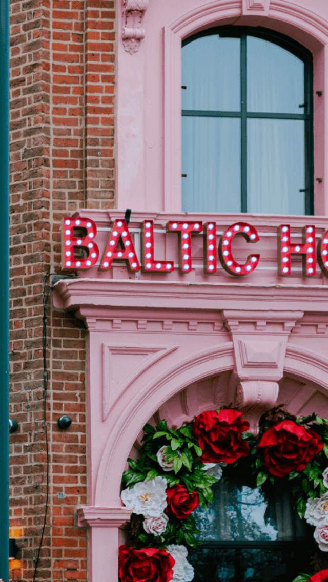 The exterior to an old warehouse building that has been turned into a hotel with a baby pink door front. The doorway has red and white roses hanging.