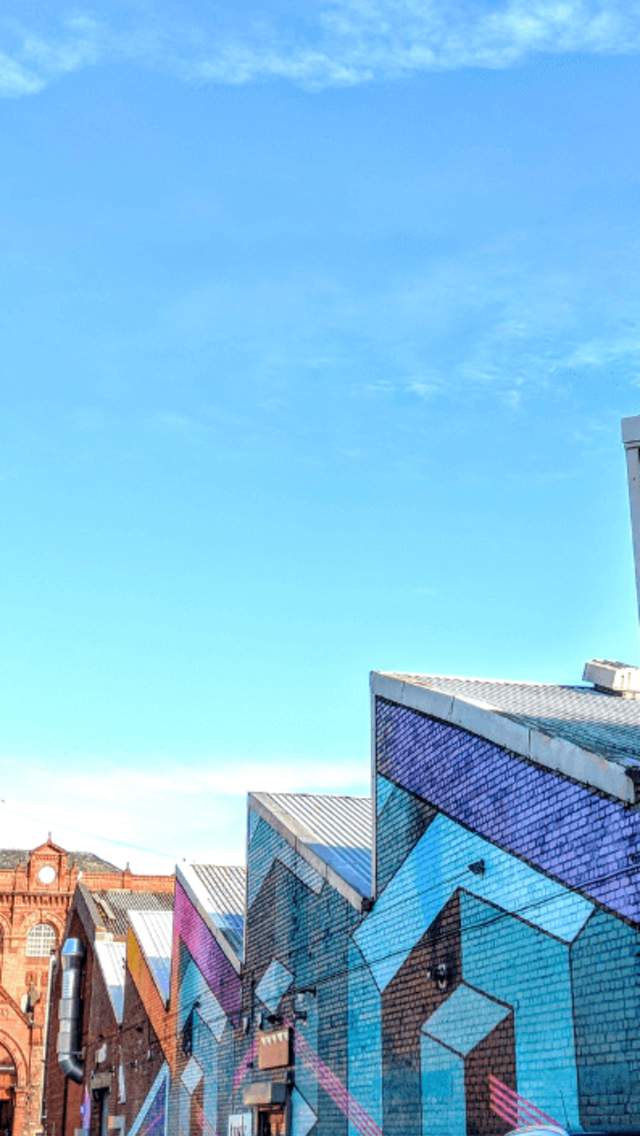 A jagged roof on a warehouse building with colorful geometric shapes painted on. Ahead is a large red-brick building which is the former Cains Brewery.