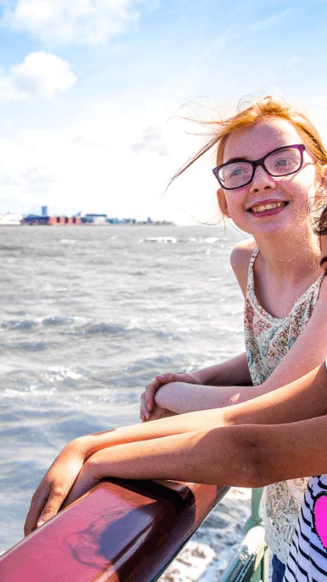 Two young girls stand on the Mersey Ferry smiling on a sunny day