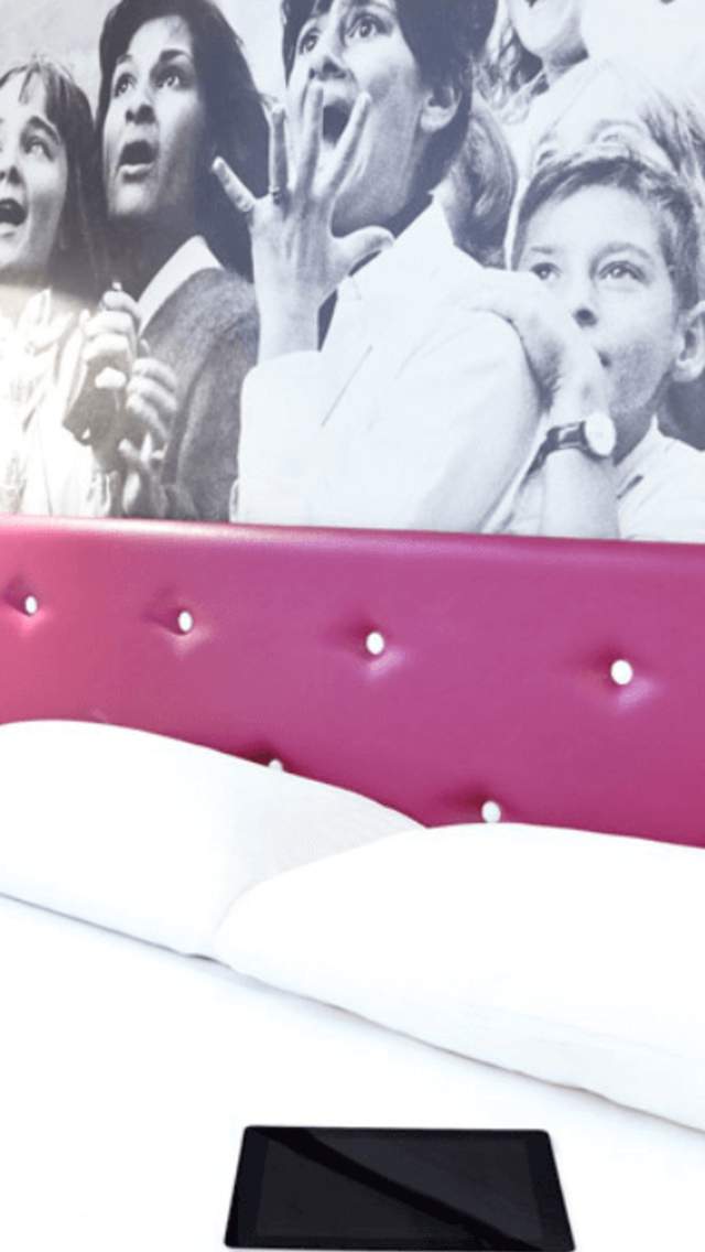 A double bed in a hotel room. There are white bed sheets and a pink, leather-look headboard. Behind the bed is a black and white photo of an excited crowd.