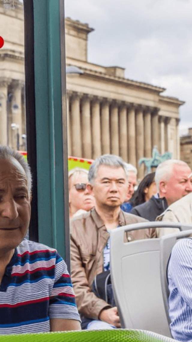 A group of people on the top deck of an open top bus tour of Liverpool. In the background the large neo-classical St George's Hall building can be seenn