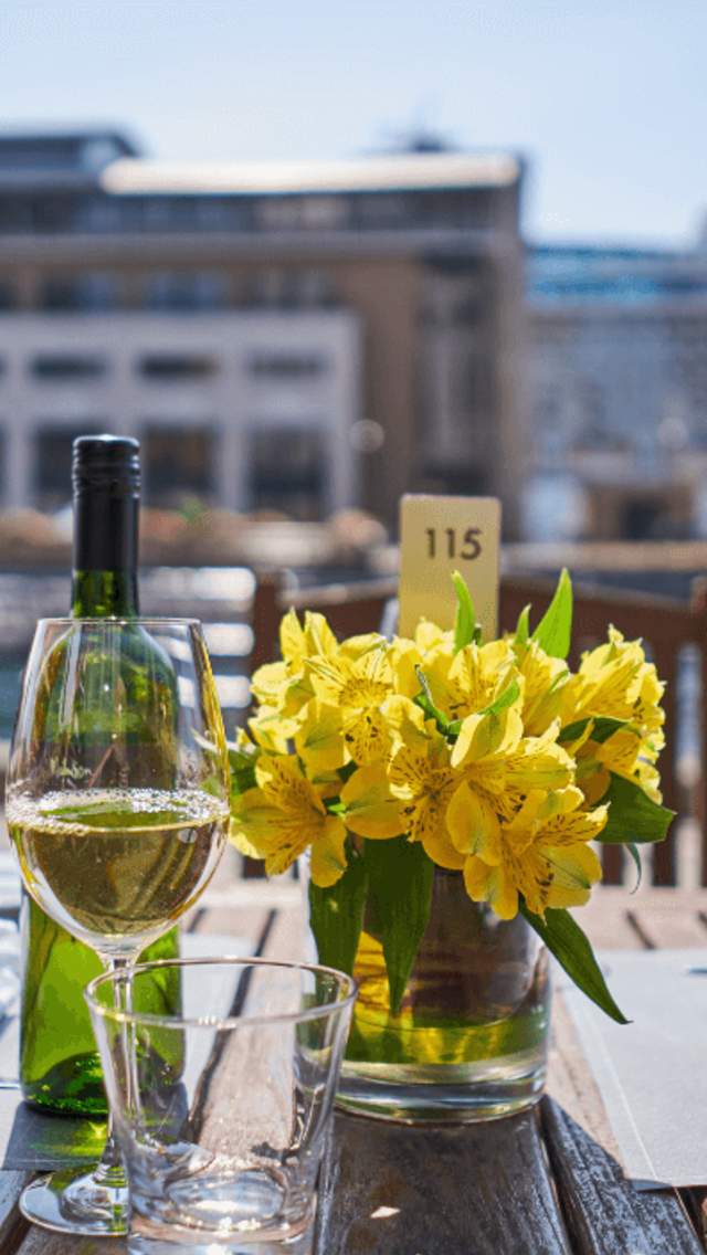 A table alongside one of the Liverpool docks, outside the Malmaison Hotel. It is a wooden outdoor table with a bunch of yellow flowers on the middle of the table, a bottle of white wine and table settings.