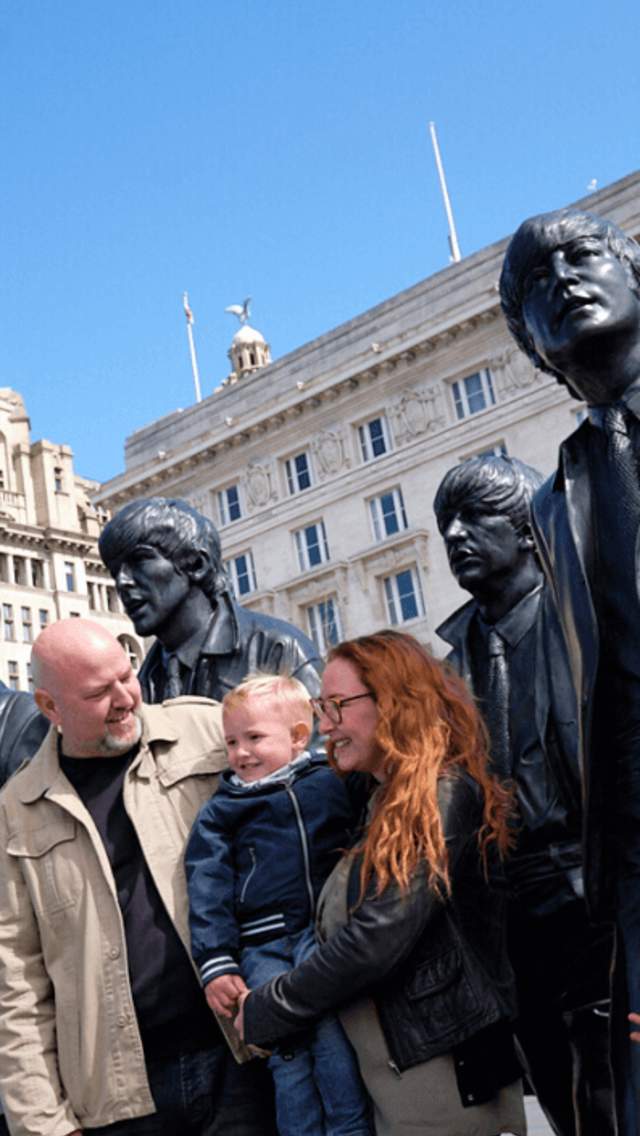 A family standing smiling next to the Beatles Statue and the Cunard and Liver Building on a sunny day.