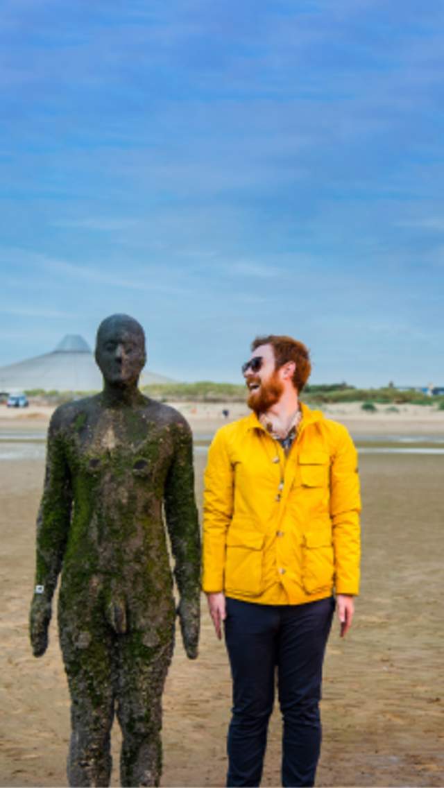 A man in a yellow rain coat stands next to a metal statue on Crosby Beach