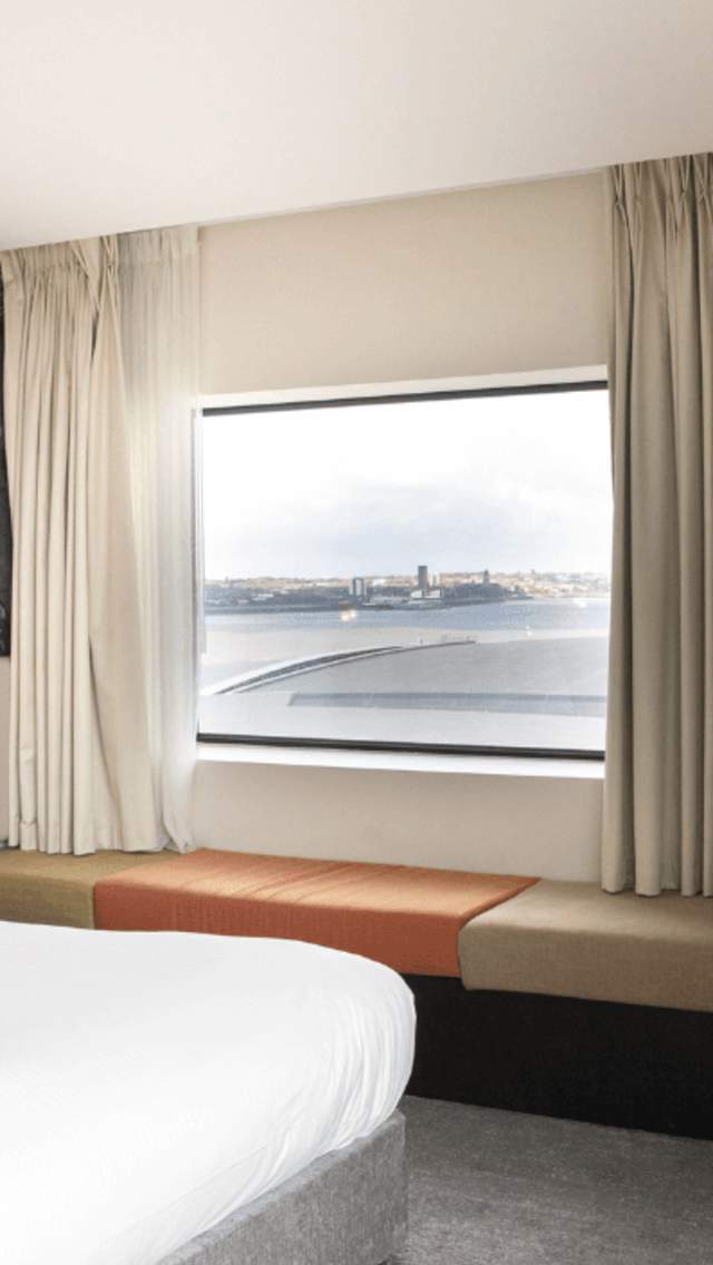 A light, spacious hotel room with a large bed with white sheets. Behind the bed is a black and white historic image of Liverpool Docklands. There's a cushioned bench style seat underneath a window, with beige curtains open.