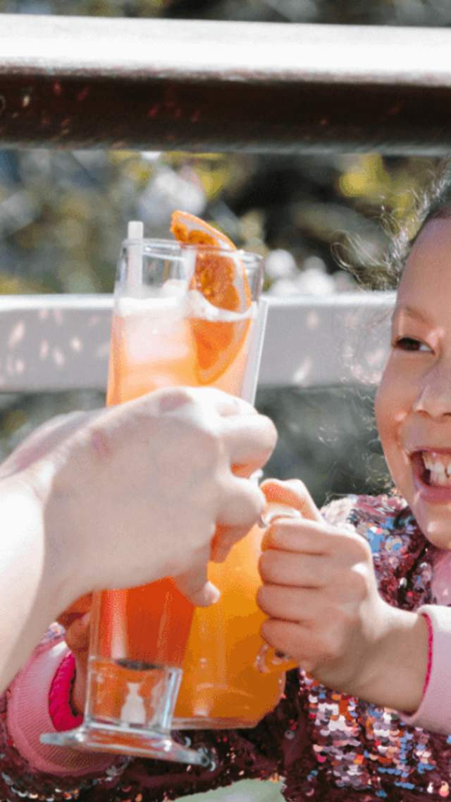 A young girl is wearing a pink sequinned jacket, her hair is in a high pony on top of her head with a colourful hair clip. She is holding a glass with an orange coloured glass and cheersing it to an adult who has a tall glass with colourful drink garnished with fruit.