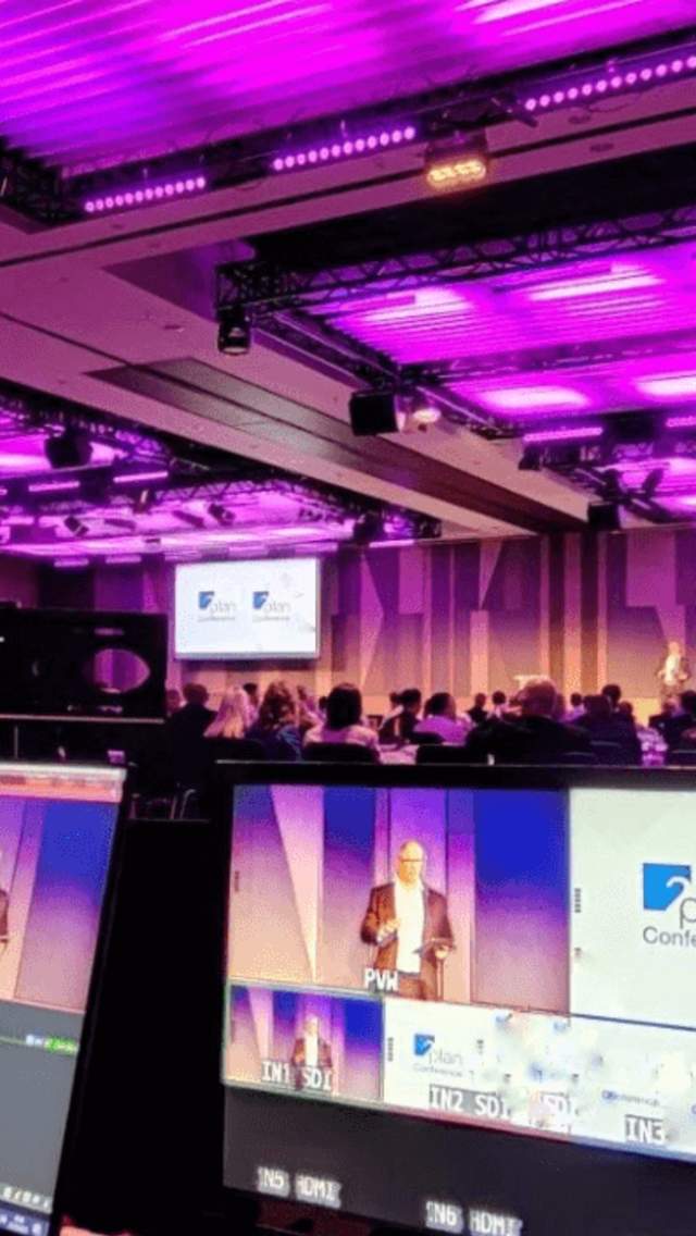 Inside a hotel conferencing room, the room is draped and lit with pink neon lights. There is a speaker on stage and the image is taken from the point of view of someone behind an AV desk with screens showing the event being broadcasted.