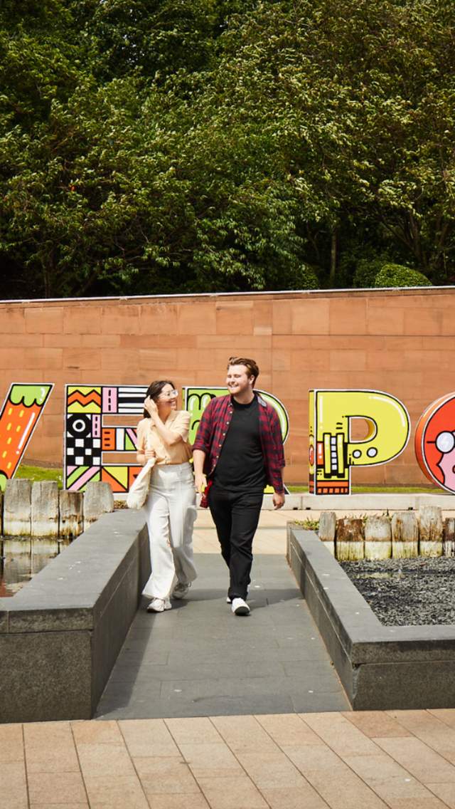 A young female and male walk happily. In the background there is a large 3D letter sign that reads 'Liverpool' and has doodle style colourful drawings decorated on each letter.