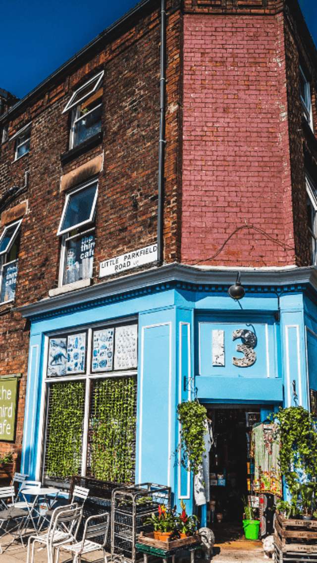 An exterior shot of a Victorian Style corner building with small sash windows and pastel blue shop front below. There are lots of plants decorating the shop front with tables and chairs outside on a sunny day.