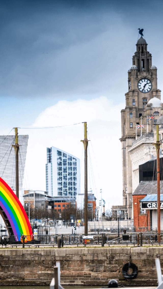A cross section image looking across towards Liverpool Pier Head. It is an Autumn day with a greyish, cloudy sky. A giant rainbow ads colour alongside the modern Museum of Liverpool building which is contrasted by the Royal Liver Building and Port of Liverpool Building.
