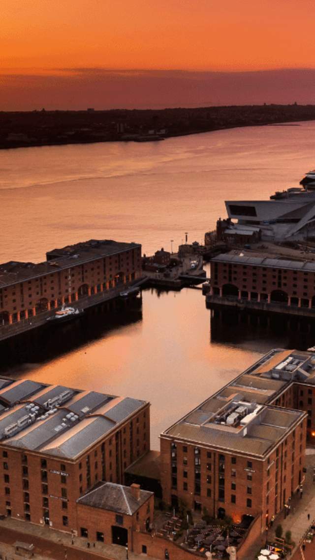 An image taken on a drone overlooking the Royal Albert Dock warehouse buildings and Liverpool Waterfront. The sun is setting on the other side of the river and the sky is a burnt orange colour.
