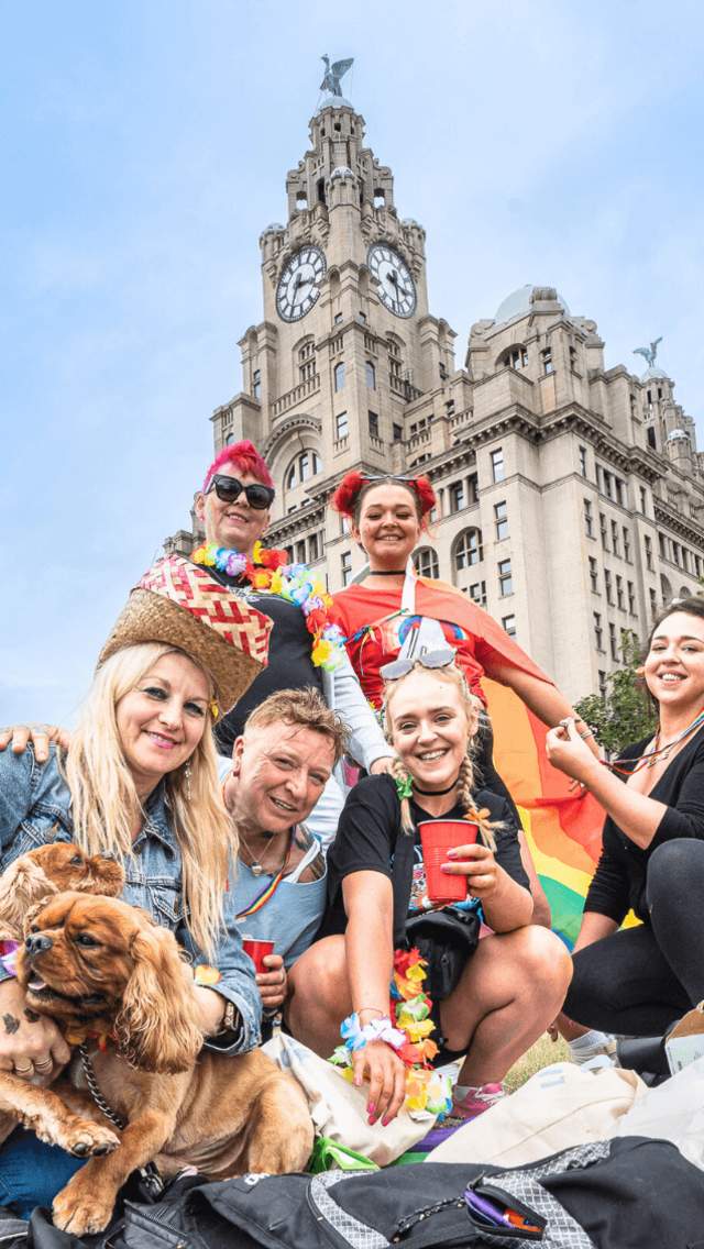 A crowd of people dressed in Pride flags and accessories on Liverpool Pier Head in front of the Liver Building