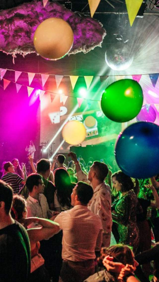 People in a warehouse with neon lights, balloons, bunting a giant disco ball and people dancing on tables.