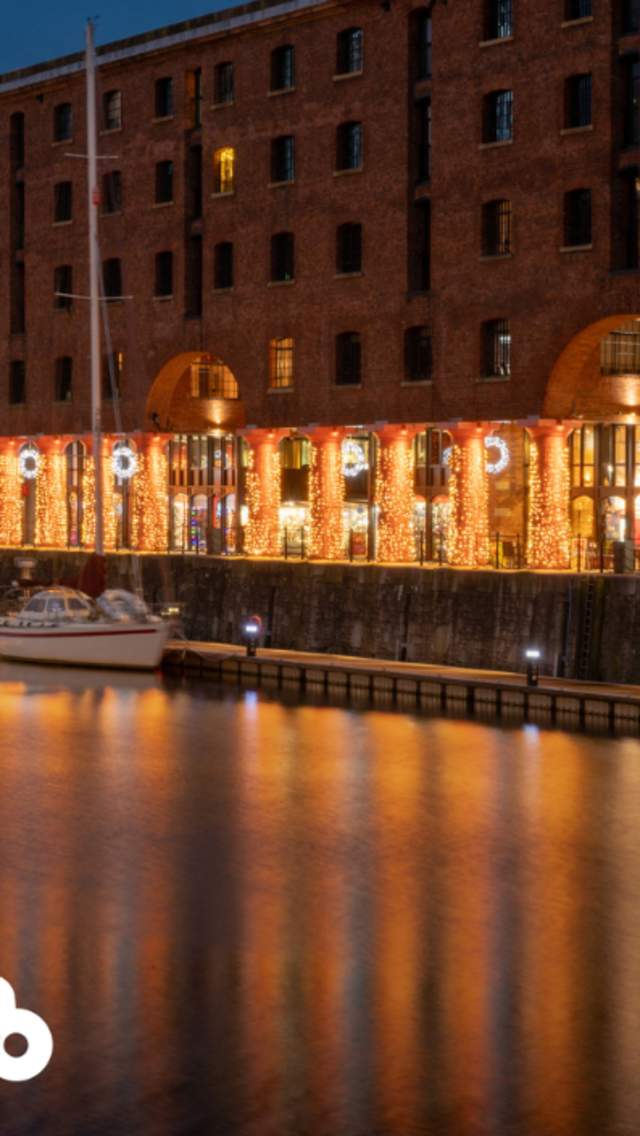 The Royal Albert Dock warehouse buildings at night tine. There are archways with large, red columns which are decorated in fairy lights.