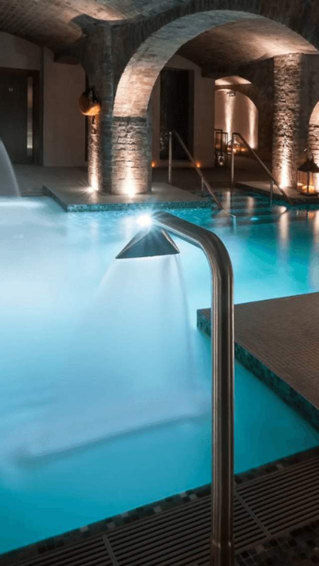 A spa pool inside a vaulted, brick space with low lighting and lanterns. The pool is blue and has hydro taps pouring into the pool.