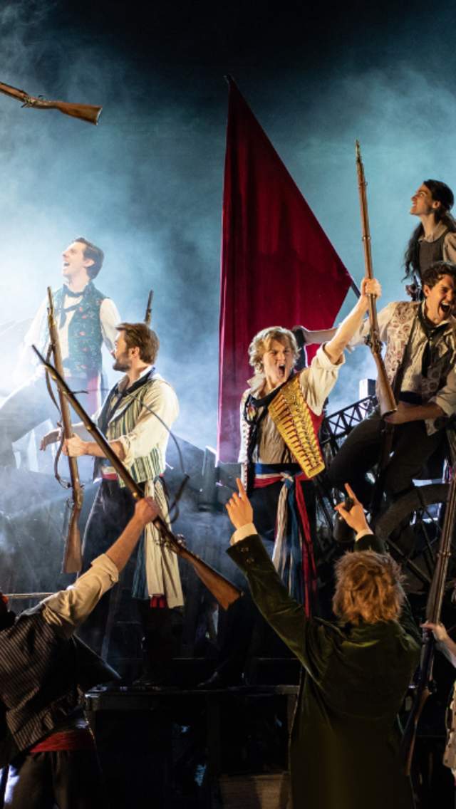 A company from the Les Miserables stage show perform at the Empire Theatre. The dramatic scene sees several actors on stage singing strongly with beams of light shining on them.