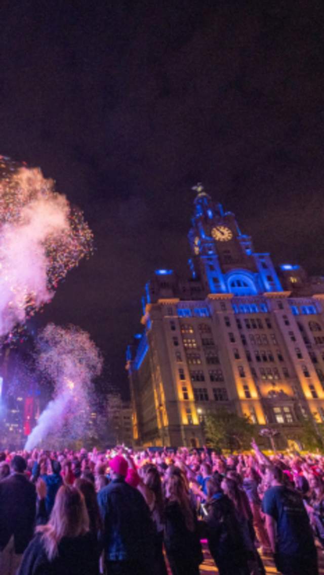 A gig on Liverpool Pier Head in front of the Liver Building at night. There are crowds and confetti cannons going off.