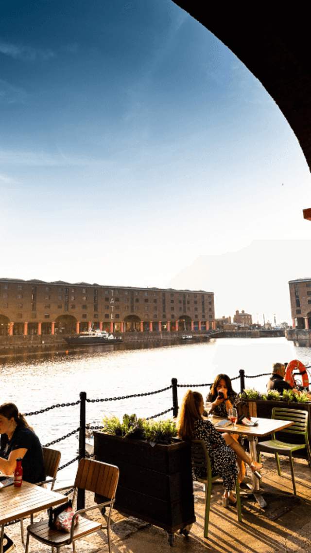 People are sitting quayside at the Royal Albert Dock on a cobbled floor at tables from a nearby restaurant. There are several warehouse buildings across a dock of water and the sun is shining over the building