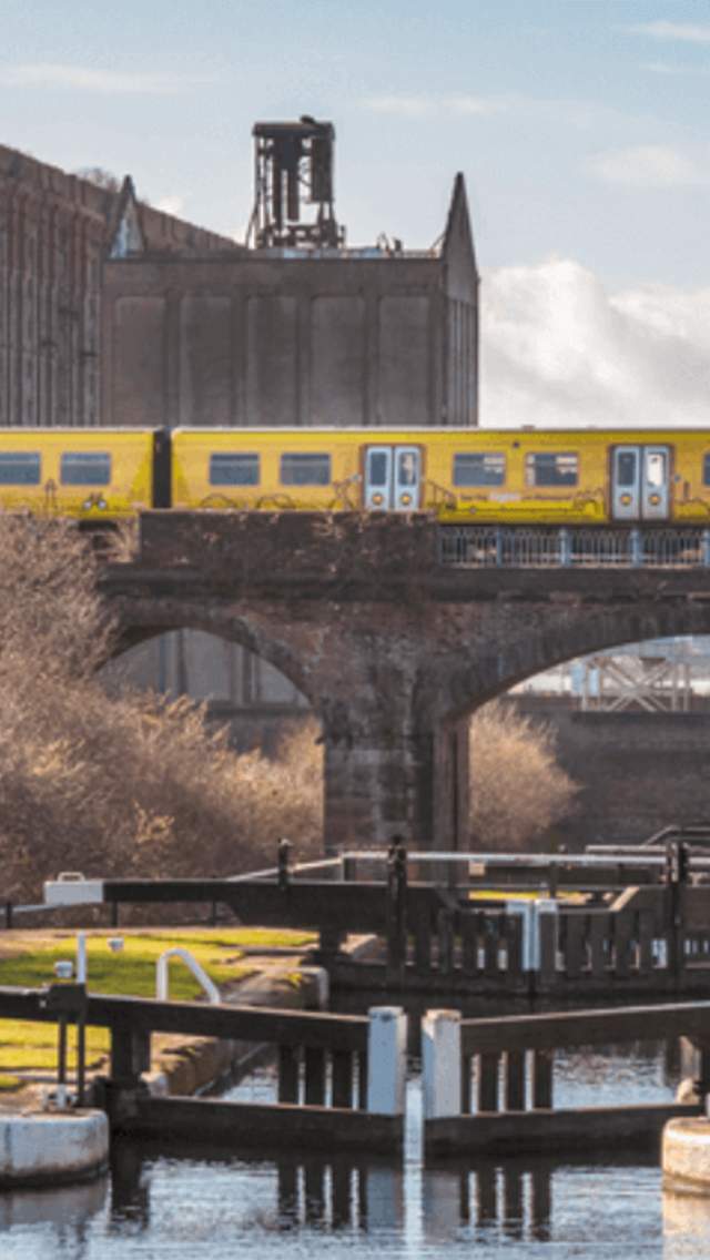A yellow train pictured along a brick railway bridge running above a canal lock system.