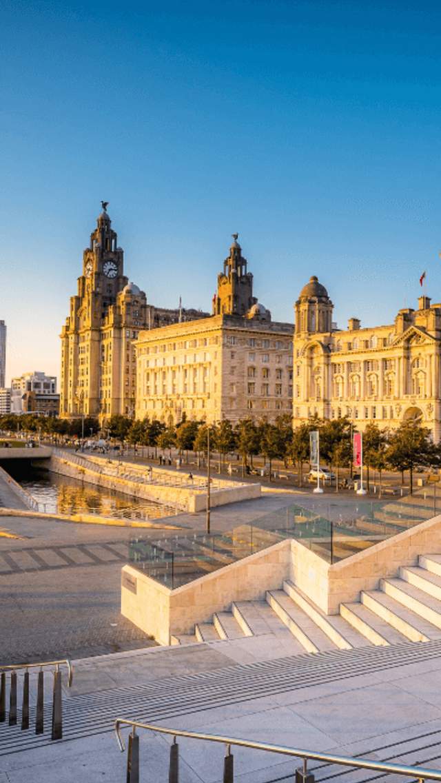 Liverpool Pier Head at golden hour. The sky is very blue and the golden sunlight is reflecting off the Liver Building, Cunard Building and Port of Liverpool. There's a cruise ship docked