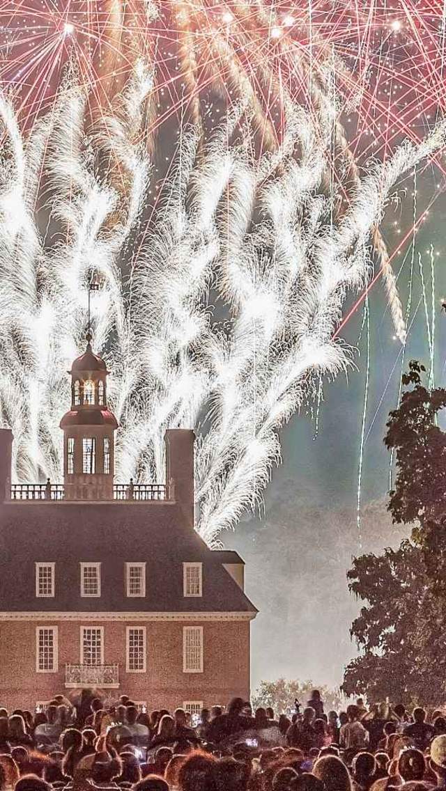 Fireworks illuminate the night sky above a historic building, surrounded by a crowd of spectators. Lush trees frame the scene, enhancing the festive atmosphere.