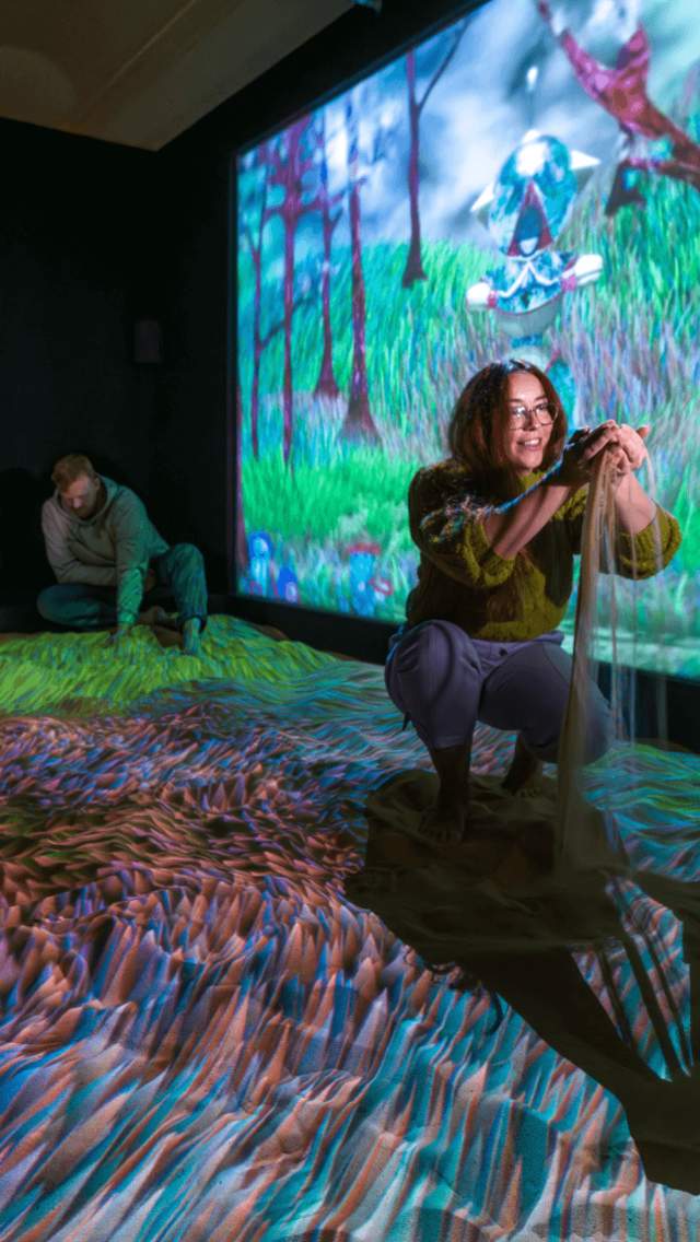 A girl picks up sand in front of a colourful projection at Subterranean Elevator