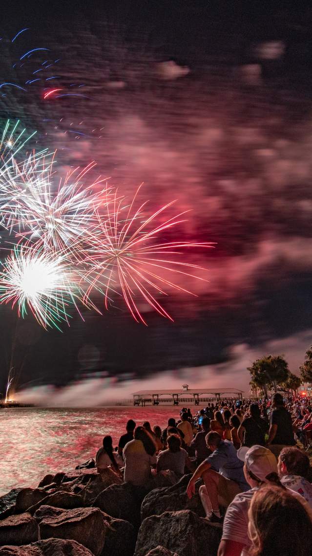 Bright fireworks burst across the sky in red, white, blue, and green over the water, reflecting across the waves below. Along the shoreline, a large crowd sits and stands on rocks and grassy areas, all facing the show. Some people are gathered in lawn chairs while others sit closer to the water’s edge, creating a relaxed but excited atmosphere.
