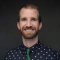 A man with a beard wearing a navy and white polka dot short with green tie.