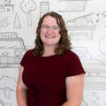 A woman with curly hair wearing a burgundy dress smiles at the camera. She stands in front of a mural that features sketches of the attractions in Kalamazoo, MI.
