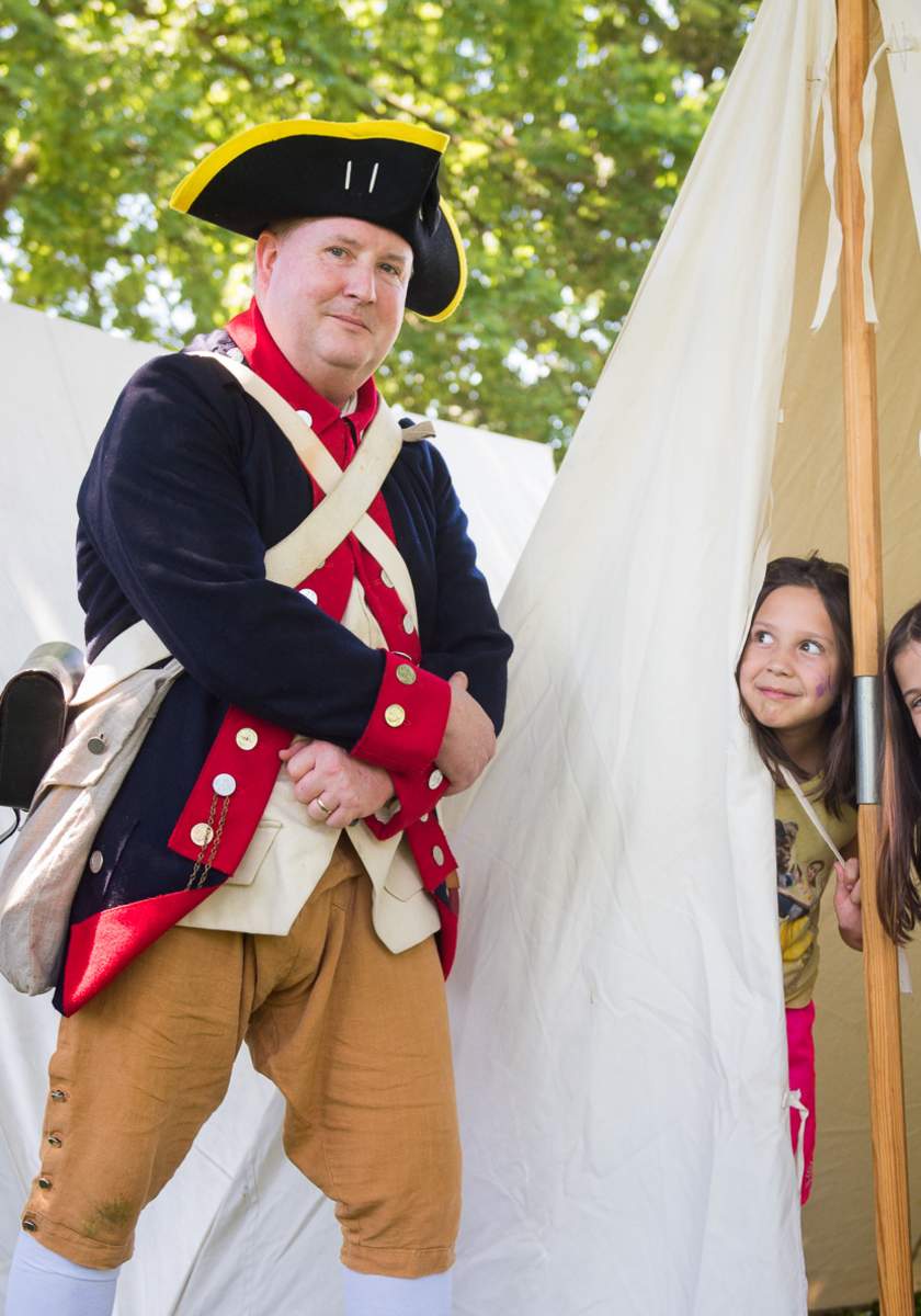 A Colonial reenactor poses in front of a tent, while two children peek out from inside