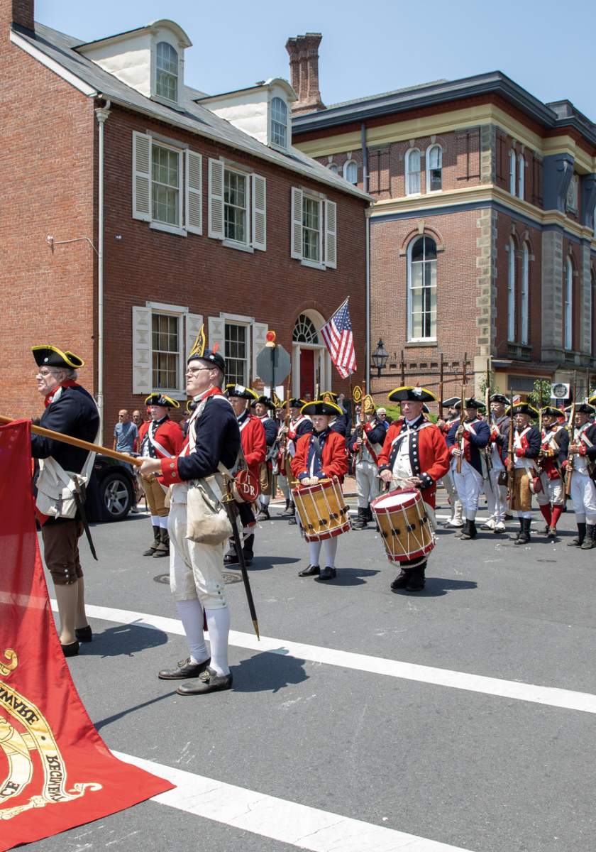 Revolutionary War reenactors, some carrying a large red British flag, march down a street lined with historic brick buildings and spectators on a sunny day.