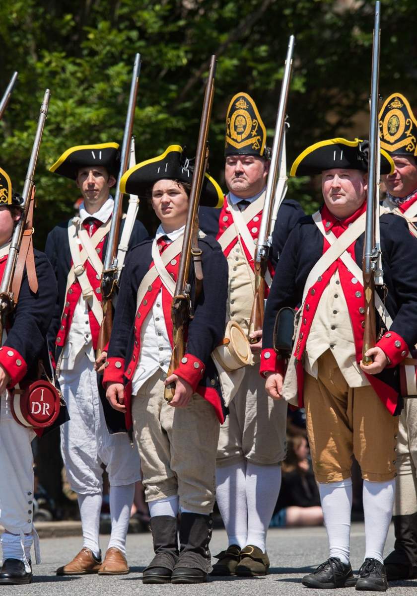 Historical reenactors in Colonial American military uniforms, holding muskets, standing in formation outdoors.