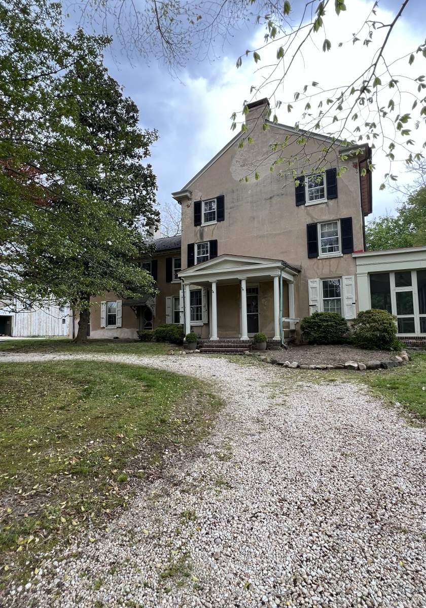 Two-story historic house with a portico entrance, set back from a gravel driveway, with a white barn visible to the left and trees around.